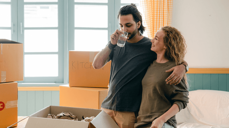 Moving boxes stacked in a new home with a couple of people with a clear glass of water in the foreground, representing the importance of testing water quality when moving into a new home