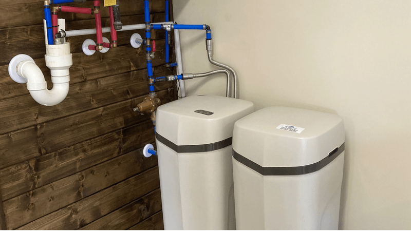 Professional whole-house water treatment system installation in a home utility room, showing a water softener and pre-filter staged neatly next to a water heater