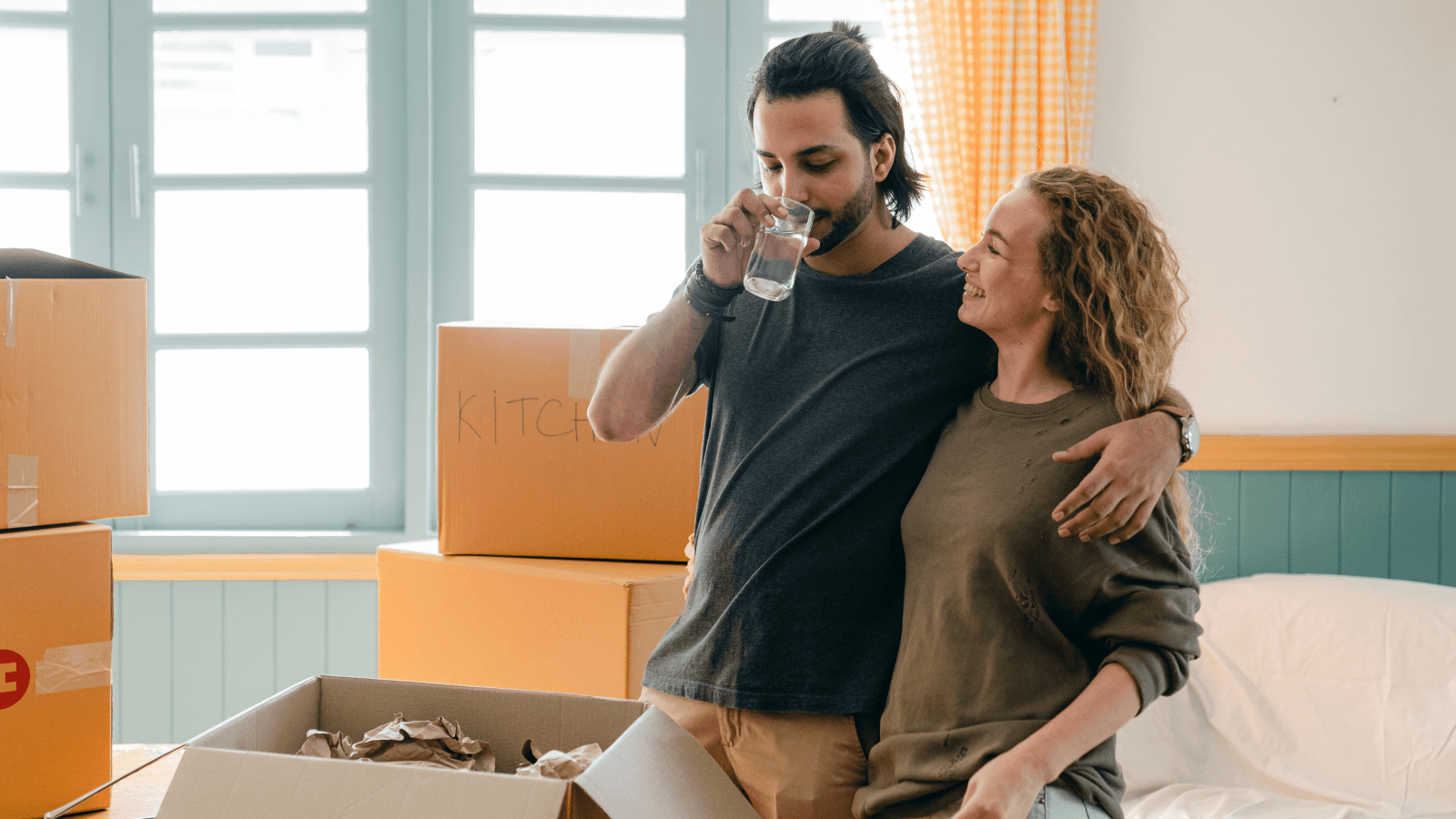 Moving boxes stacked in a new home with a couple of people with a clear glass of water in the foreground, representing the importance of testing water quality when moving into a new home